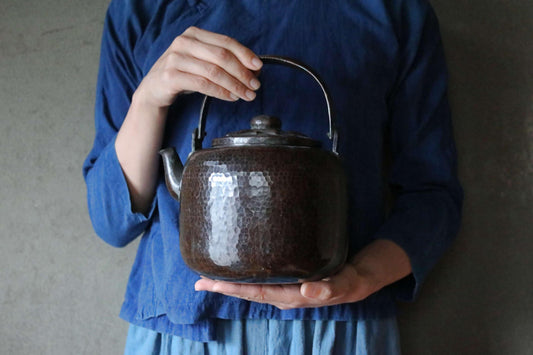 A person wearing a blue shirt is holding a dark brown, textured kettle in their hands.
