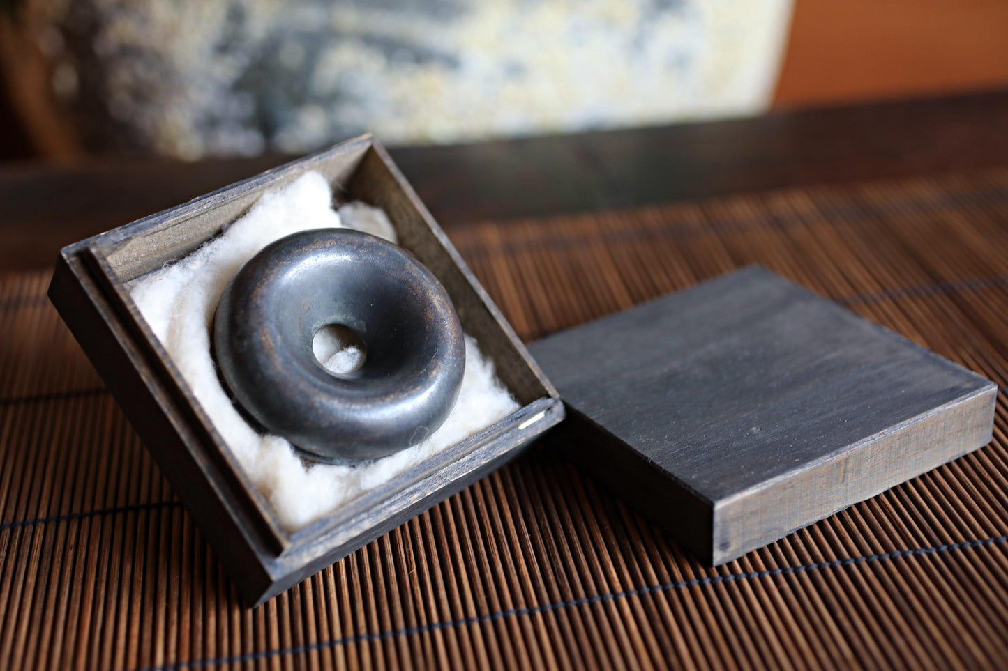 A black metal object, possibly a piece of jewelry or a decorative item, is displayed in a wooden box with a white cloth lining.