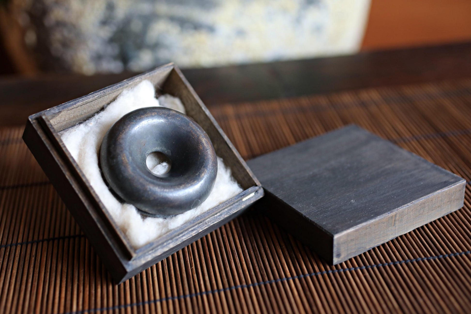 A black metal object, possibly a piece of jewelry or a decorative item, is displayed in a wooden box with a white cloth lining.