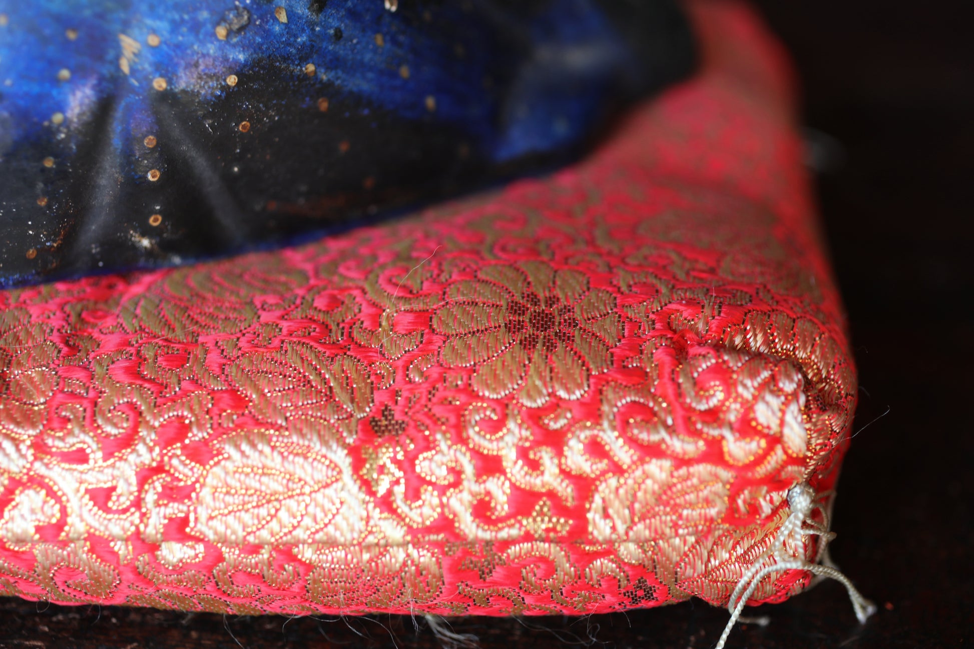 A close-up view of a red and gold patterned fabric, possibly a cushion or pillow, with a blue and black background.