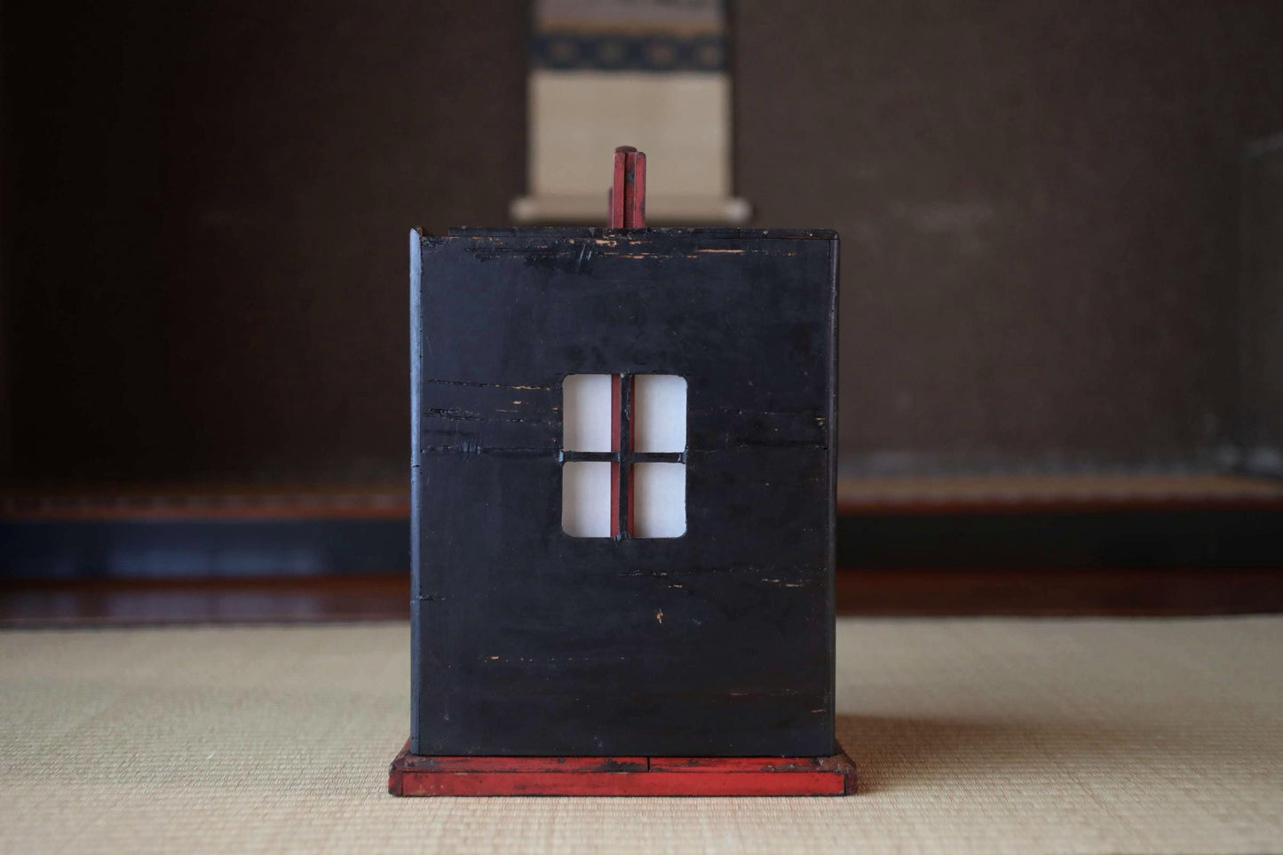 A black wooden box with a red trim, featuring a small window, is placed on a beige surface.