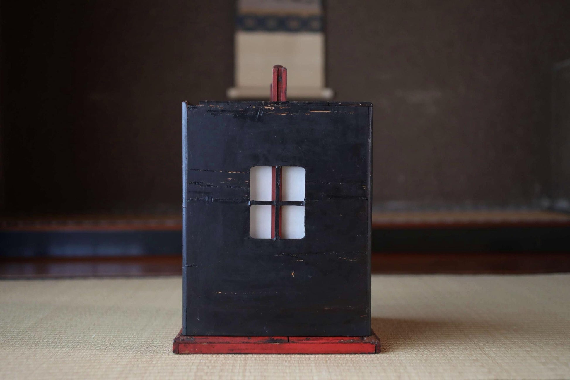 A black wooden box with a red trim, featuring a small window, is placed on a beige surface.