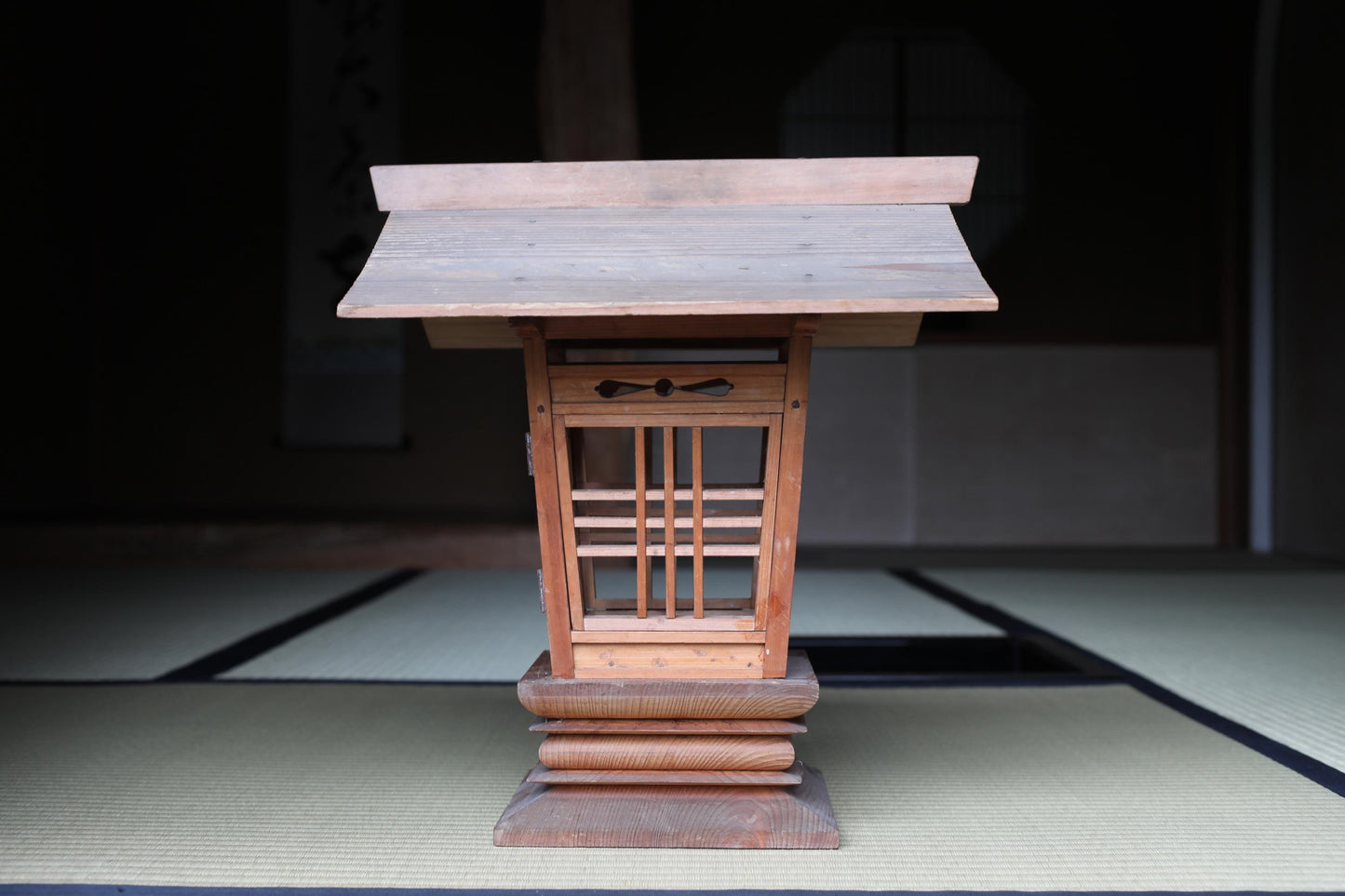 A wooden shrine or altar with a sloping roof, featuring a lattice design and a small window, situated on a tatami mat.