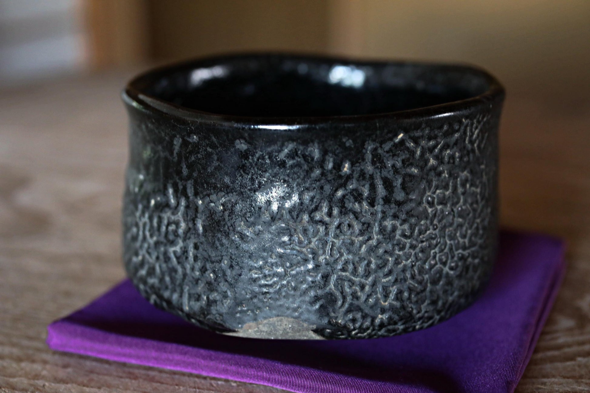 A black ceramic bowl with a textured surface sits on a purple cloth on a wooden surface.