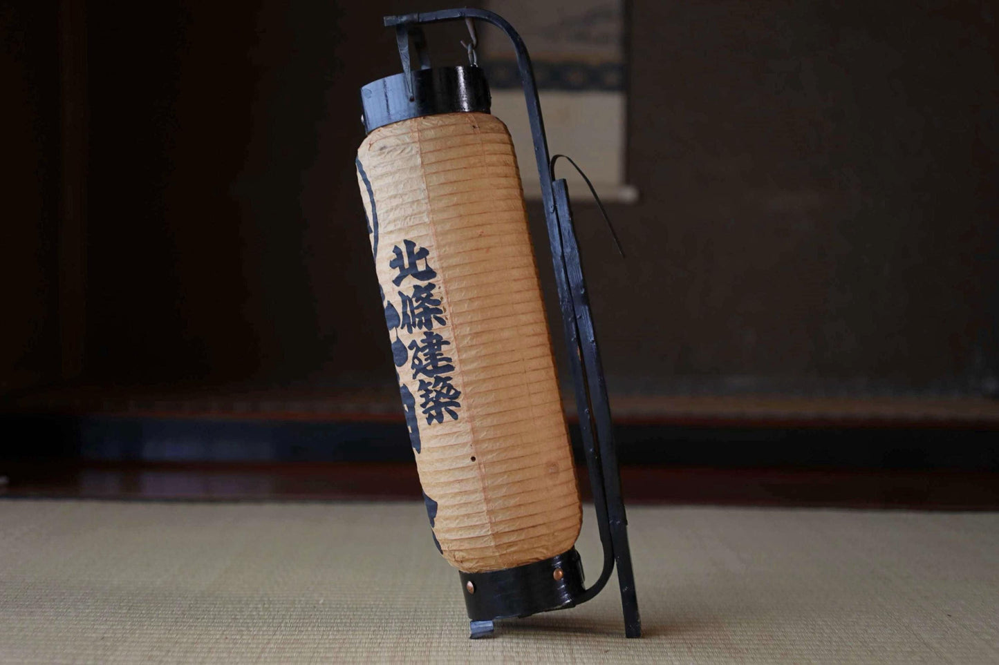 A traditional Japanese paper lantern with a black metal frame stands on a tatami mat.