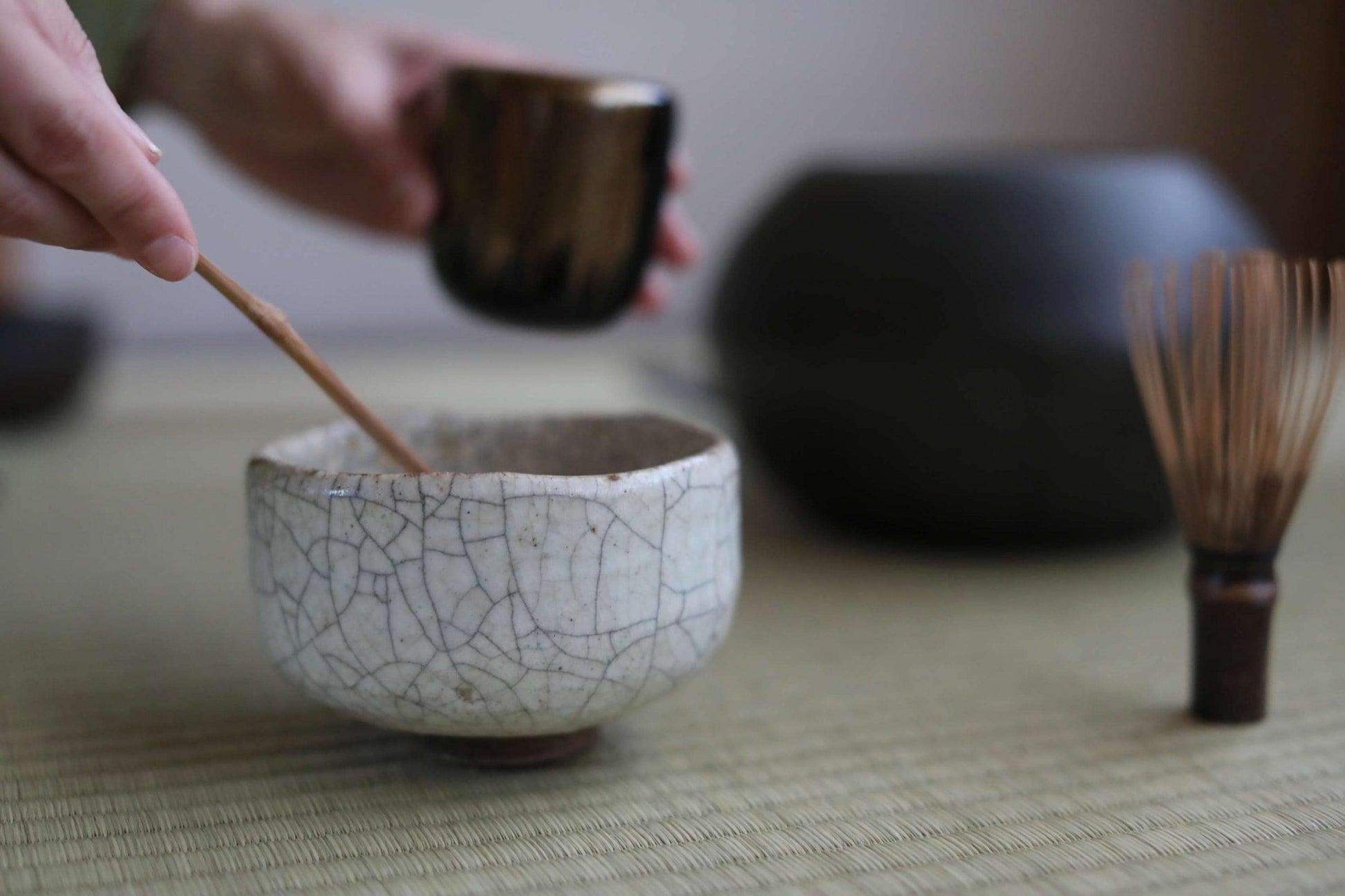 A person's hands are holding a small ceramic cup and a wooden whisk, preparing to whisk something in a bowl.