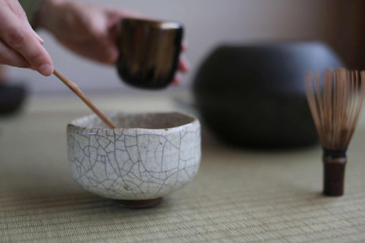 A person's hands are holding a small ceramic cup and a wooden whisk, preparing to whisk something in a bowl.