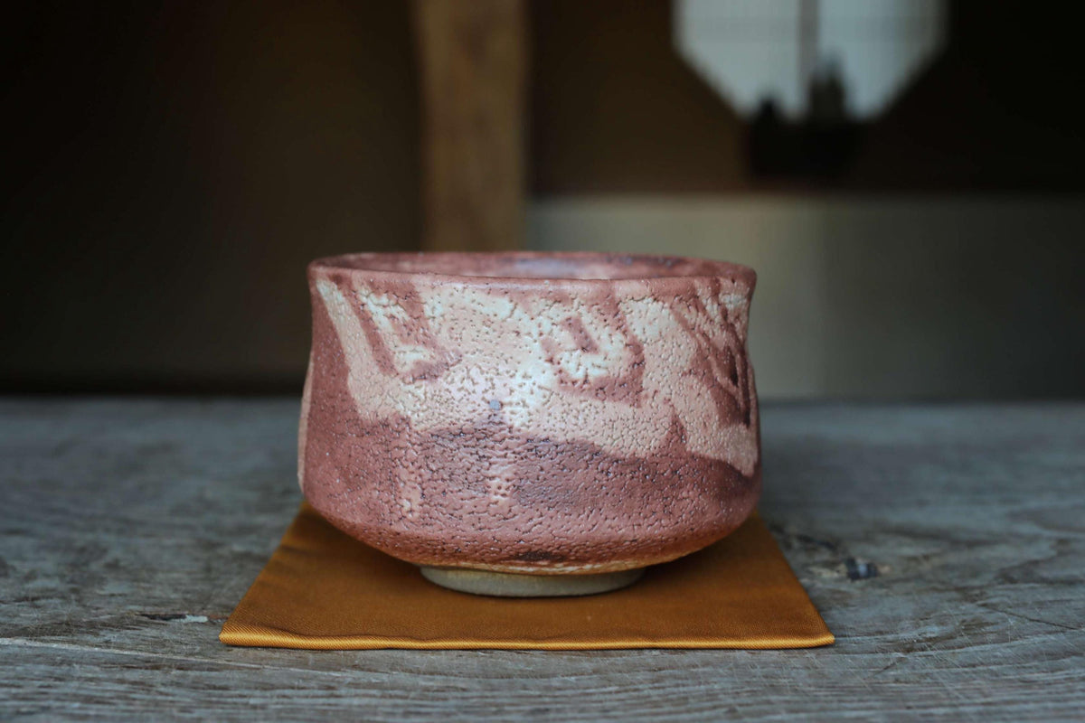 A ceramic bowl with a pinkish-brown color and a white pattern sits on a brown cloth on a wooden surface.