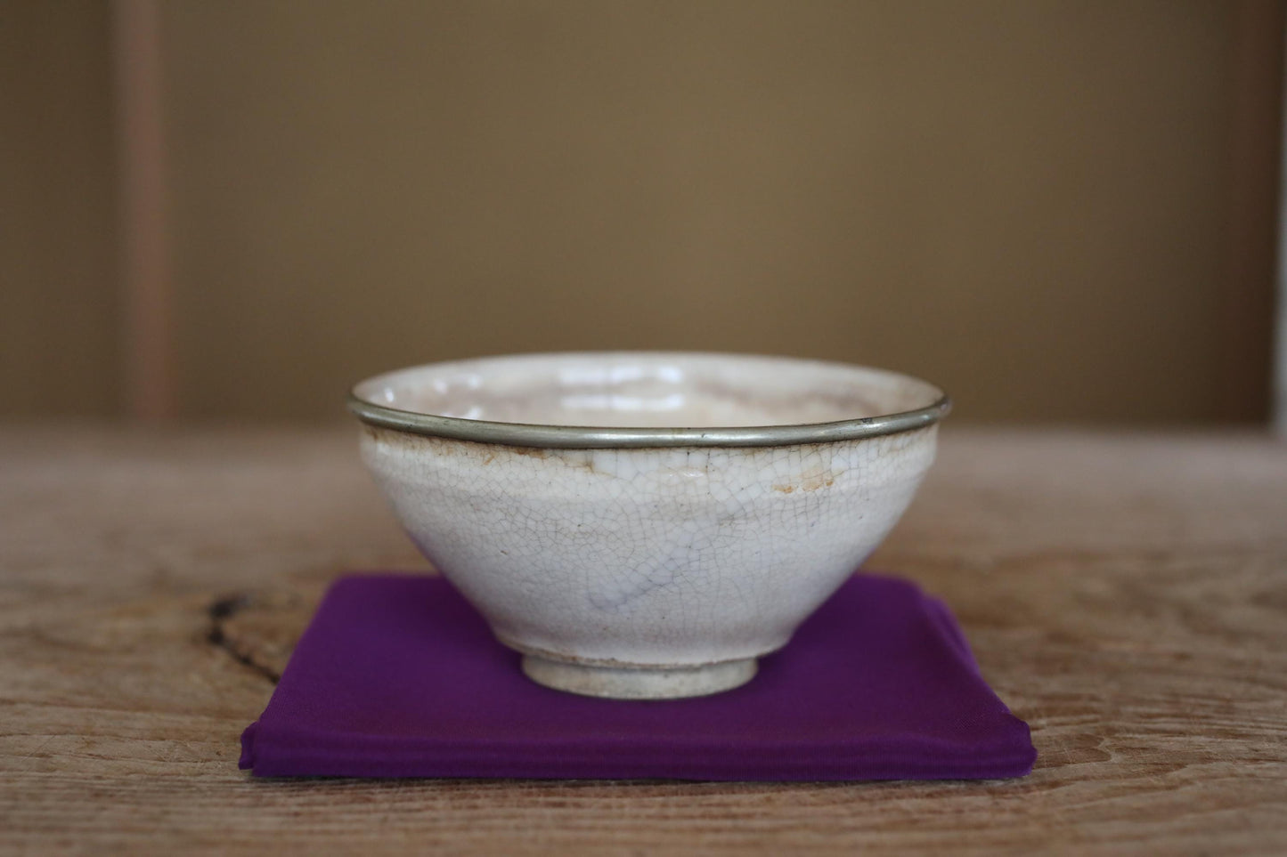 A white ceramic bowl with a silver rim sits on a purple cloth on a wooden surface.