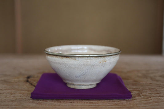 A white ceramic bowl with a silver rim sits on a purple cloth on a wooden surface.