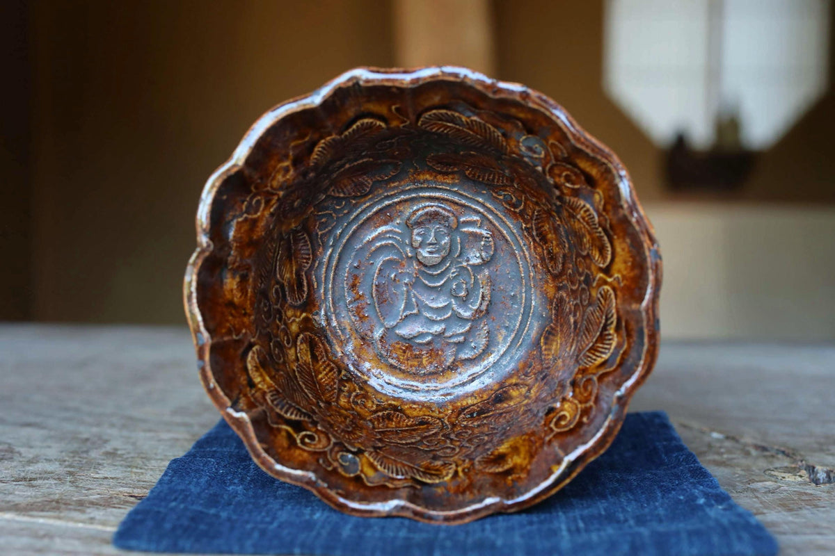 An ornate, antique-looking bowl with a carved design on the front, placed on a blue cloth.