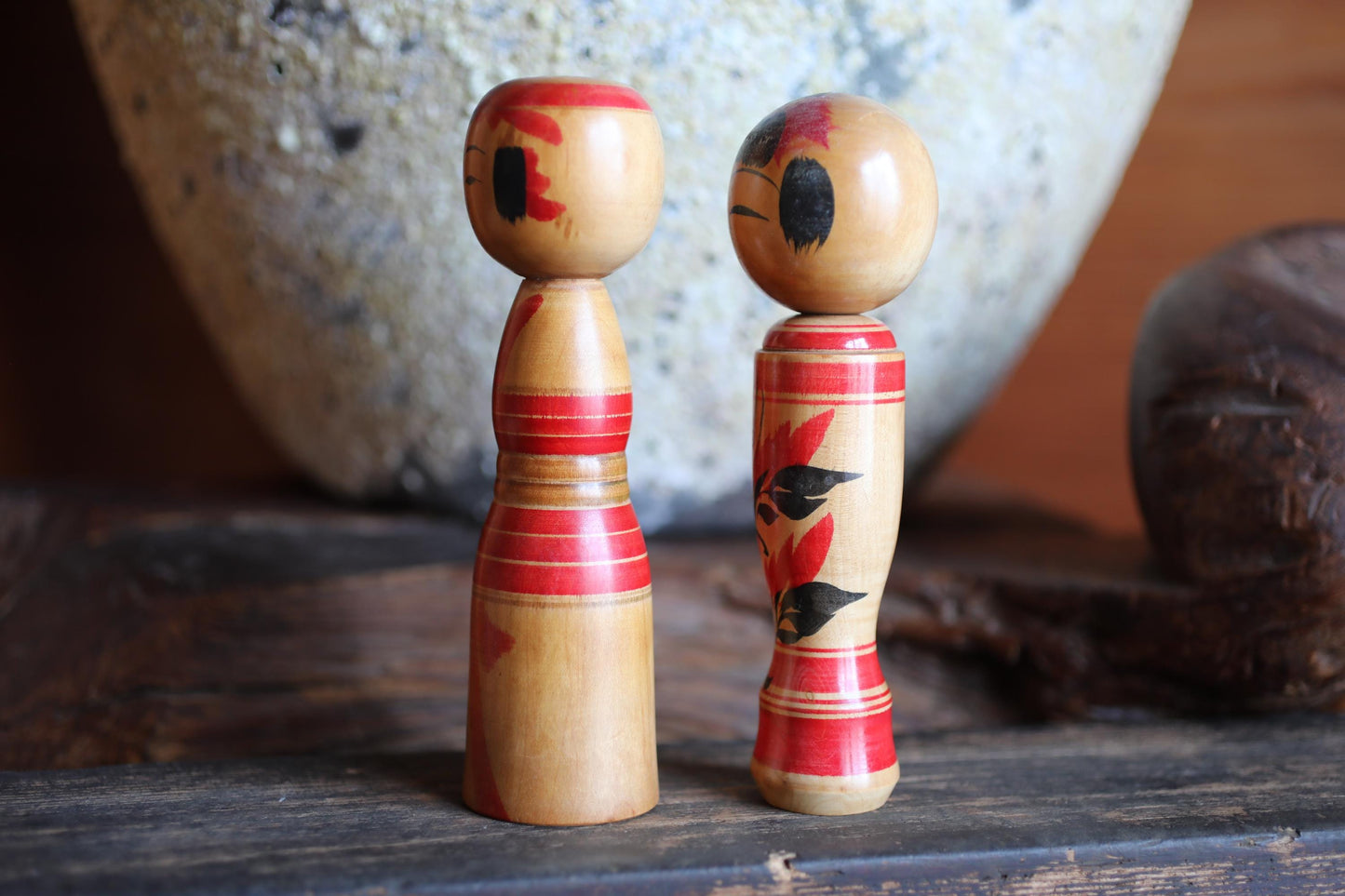 Two wooden dolls with red and black designs stand on a wooden surface, with a large stone vase in the background.