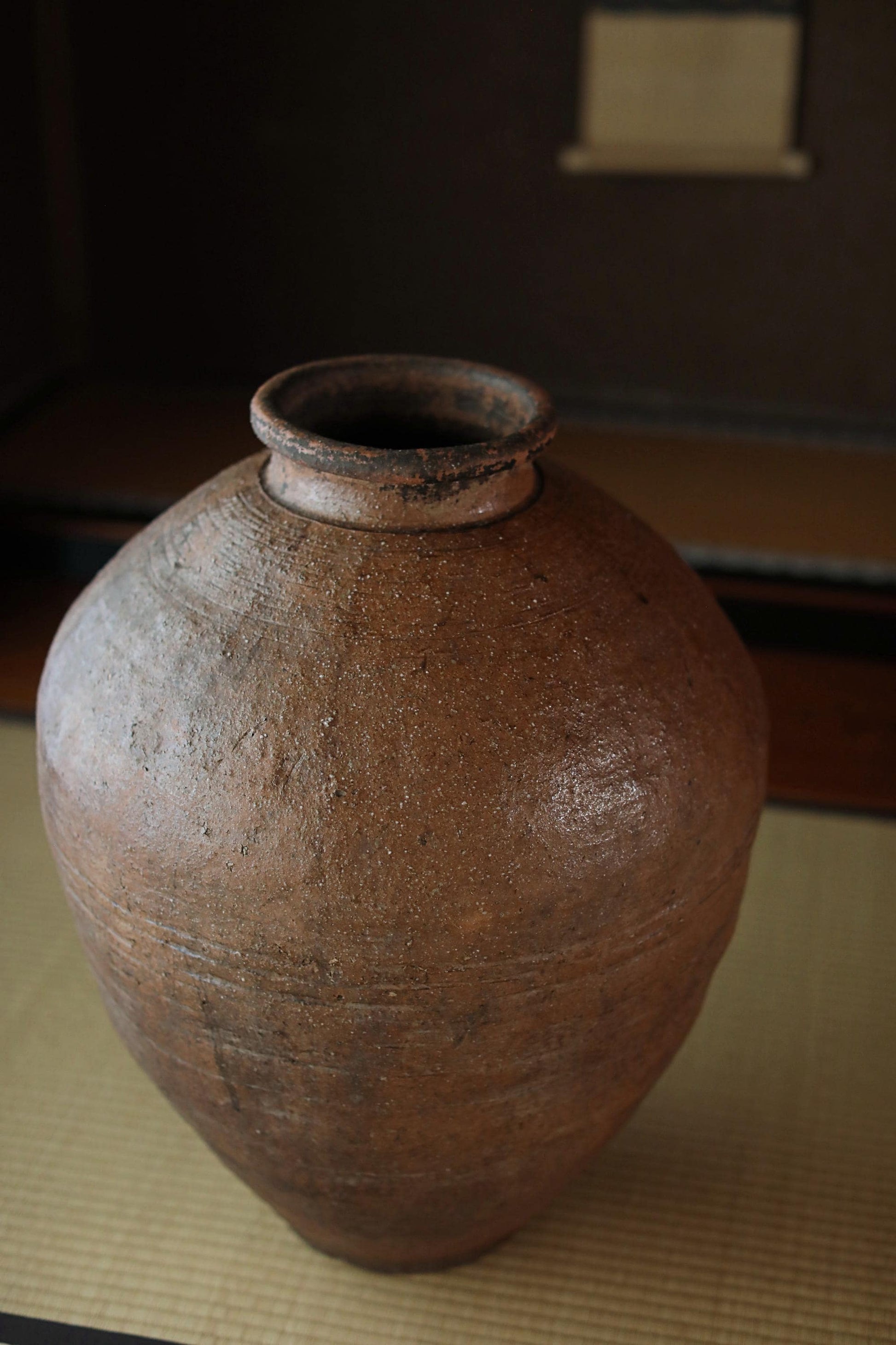 An old, large clay vase with a rusted exterior sits on a mat against a dark background.