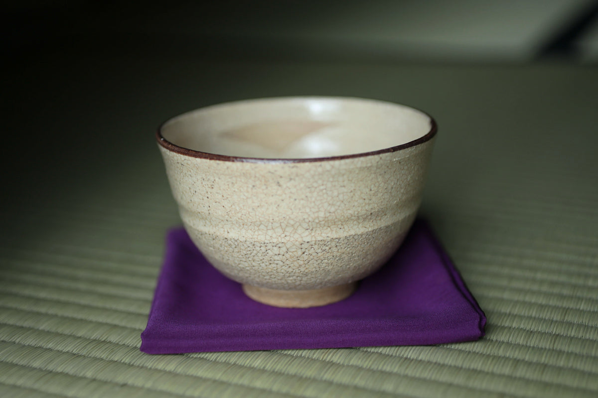 A white ceramic bowl with a brown rim sits on a purple cloth on a woven mat.
