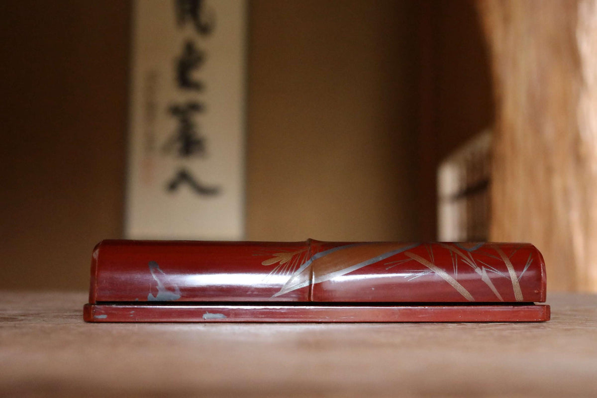 A red, wooden object with a decorative design sits on a wooden surface, with a blurred background that includes a vertical banner with Japanese characters.