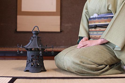 A person in a traditional Japanese kimono sits on the floor, with a small black lantern or tea lamp next to them.