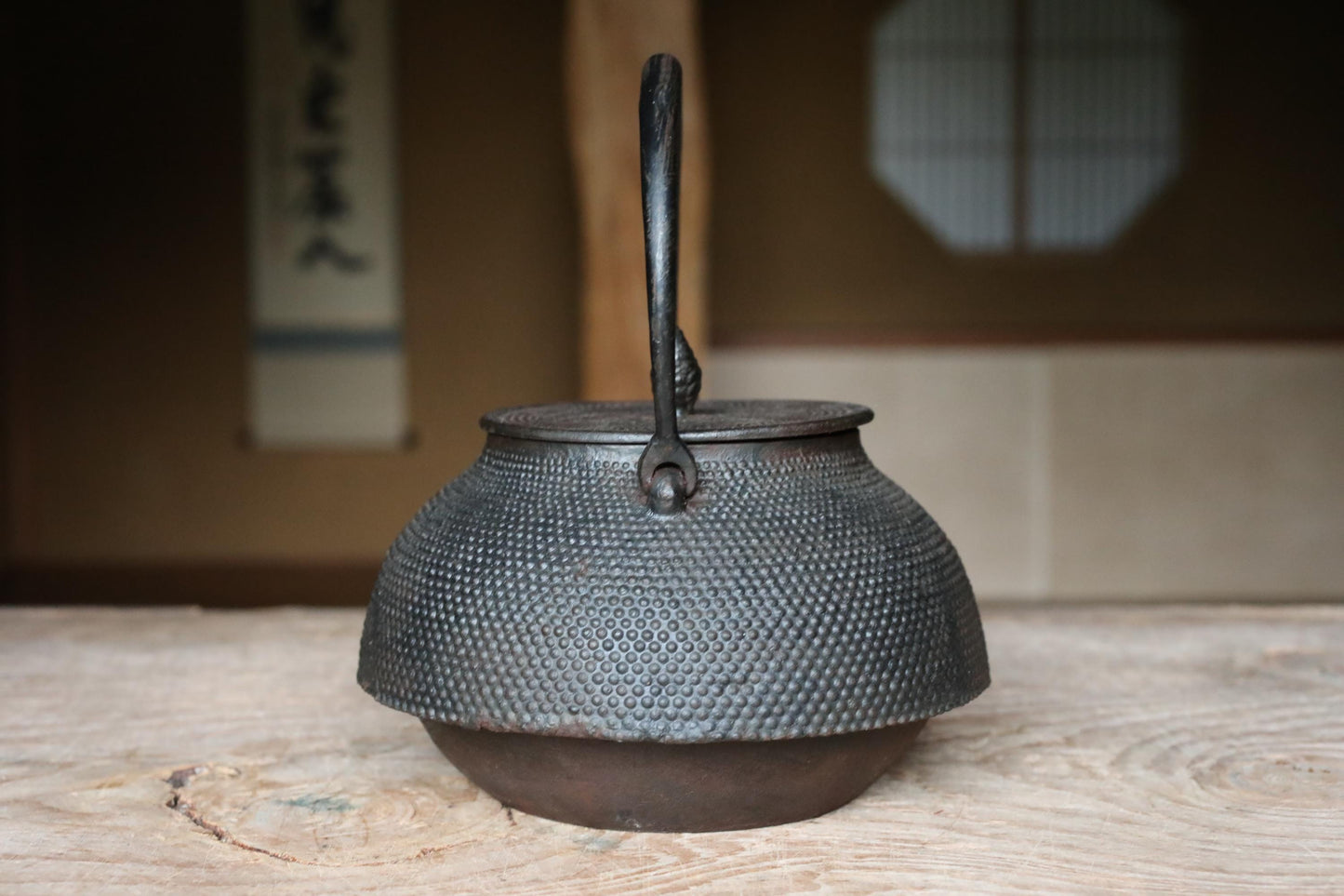 A black metal teapot with a handle and spout sits on a wooden surface, with a blurred background that includes a window and a wall with Japanese characters.