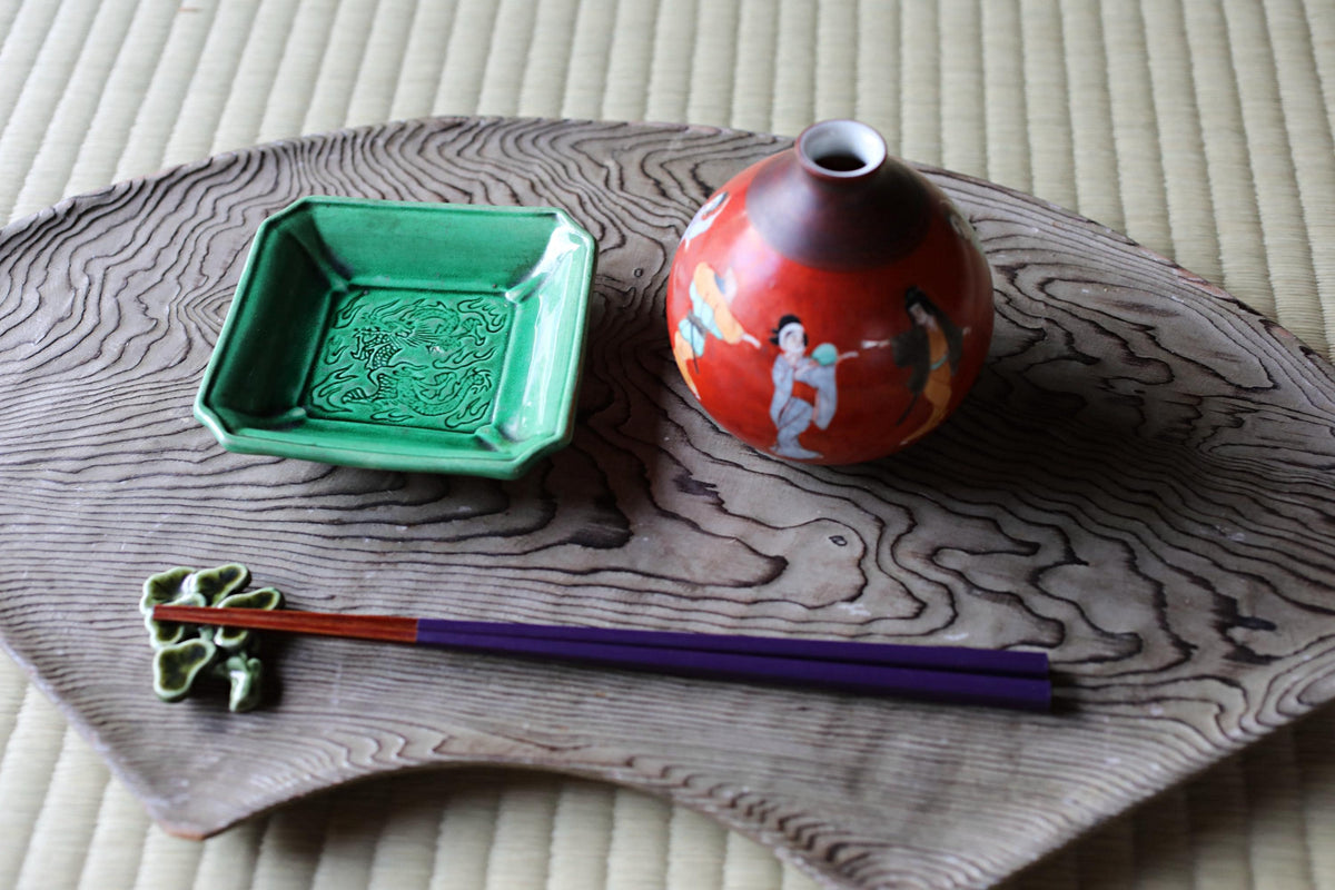 a wooden tray with various decorative items placed on it, including a red vase, a green dish, and a pair of chopsticks.