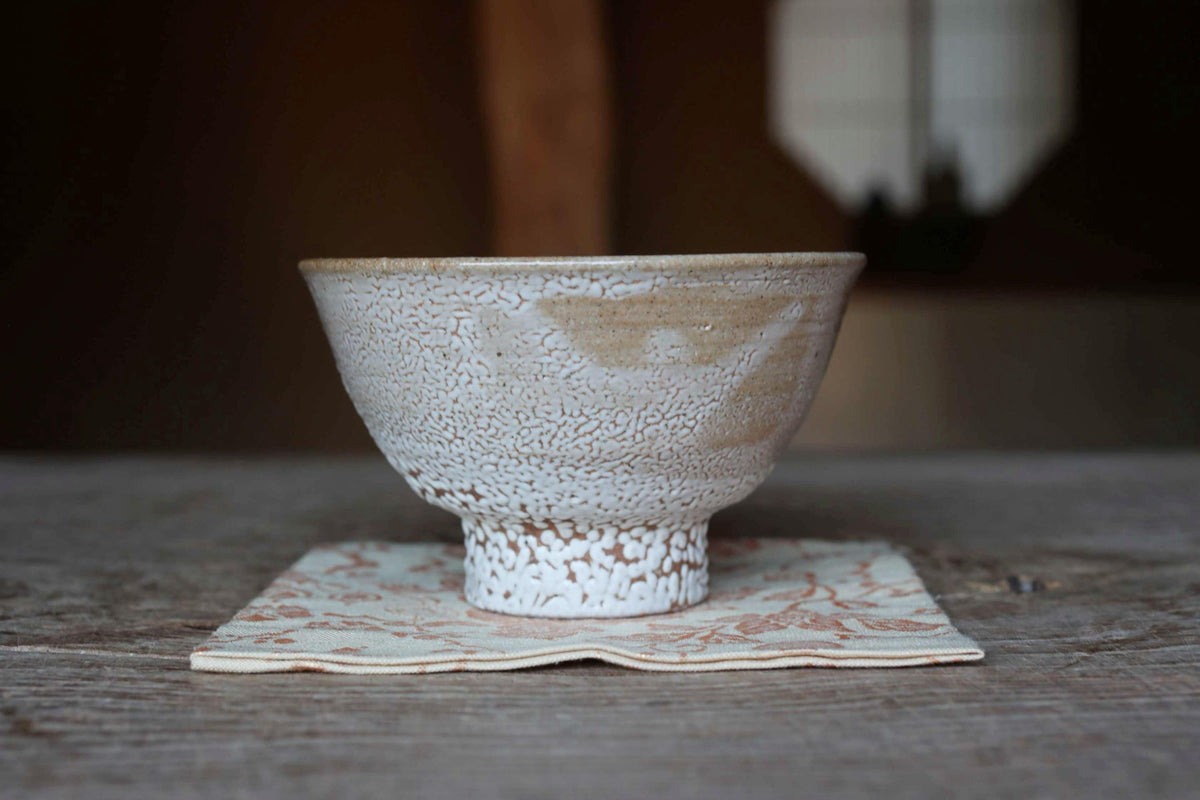 A white ceramic bowl with a speckled texture sits on a white and orange floral napkin on a wooden table.