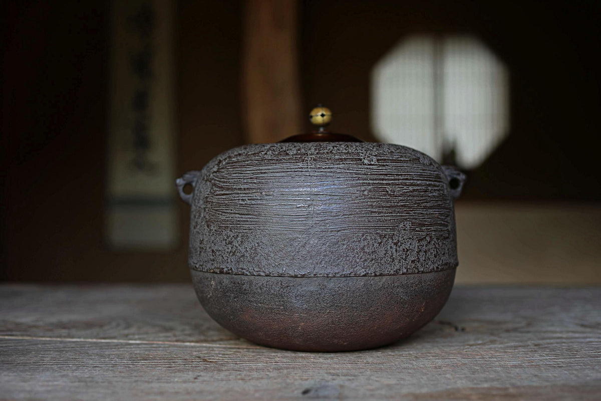 A dark brown ceramic bowl with a gold-colored lid sits on a wooden surface, with a blurred background suggesting an indoor setting.