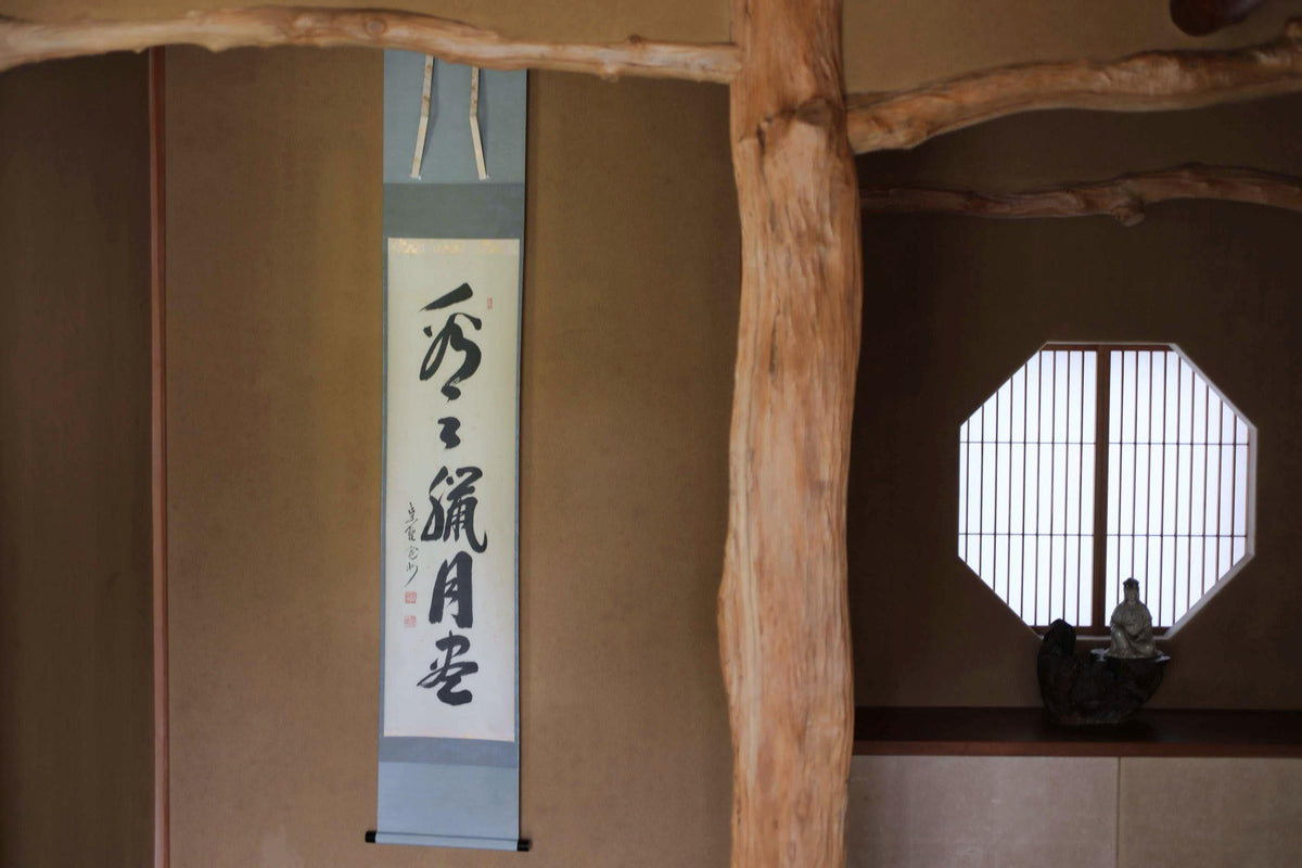 a traditional Japanese temple interior with a wooden beam and a window with a bamboo screen. A vertical banner with Japanese characters is hanging on the wall, and a small statue of a Buddha is visible in the background.