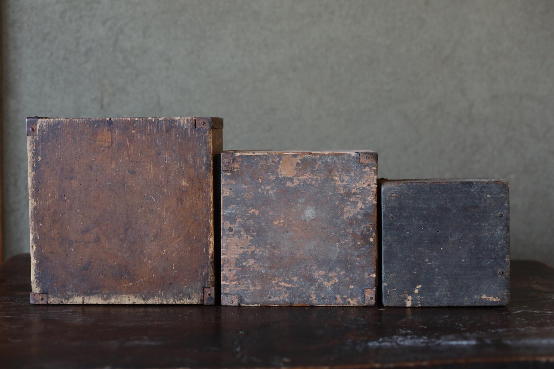 Three old, weathered wooden boxes of varying sizes are arranged on a dark surface against a gray background.