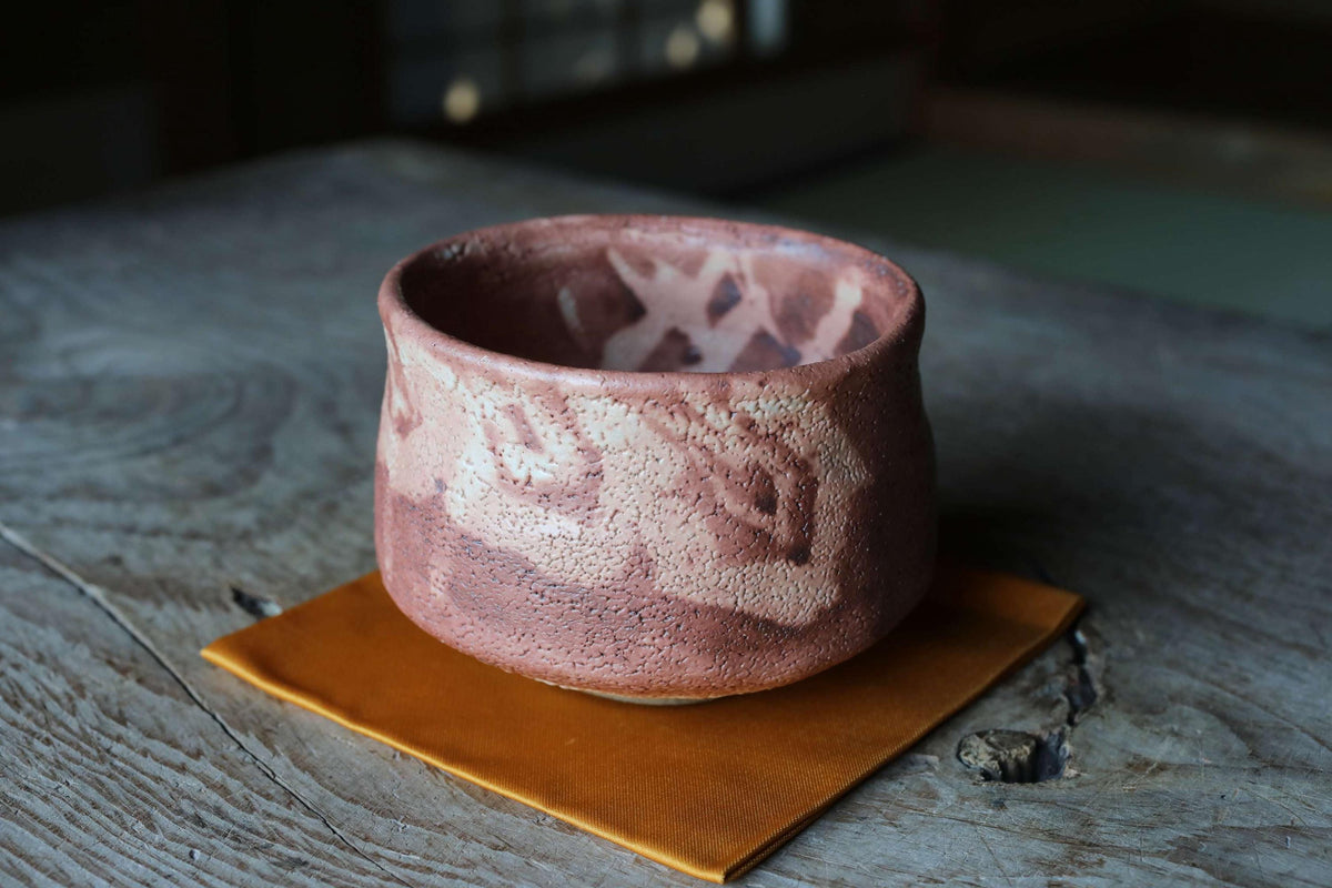 A ceramic bowl with a pinkish-brown color and a patterned design sits on a wooden surface, accompanied by a small orange cloth or mat.