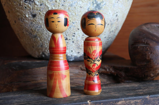 Two wooden dolls with red and yellow accents are displayed on a wooden surface, with a large stone vase in the background.