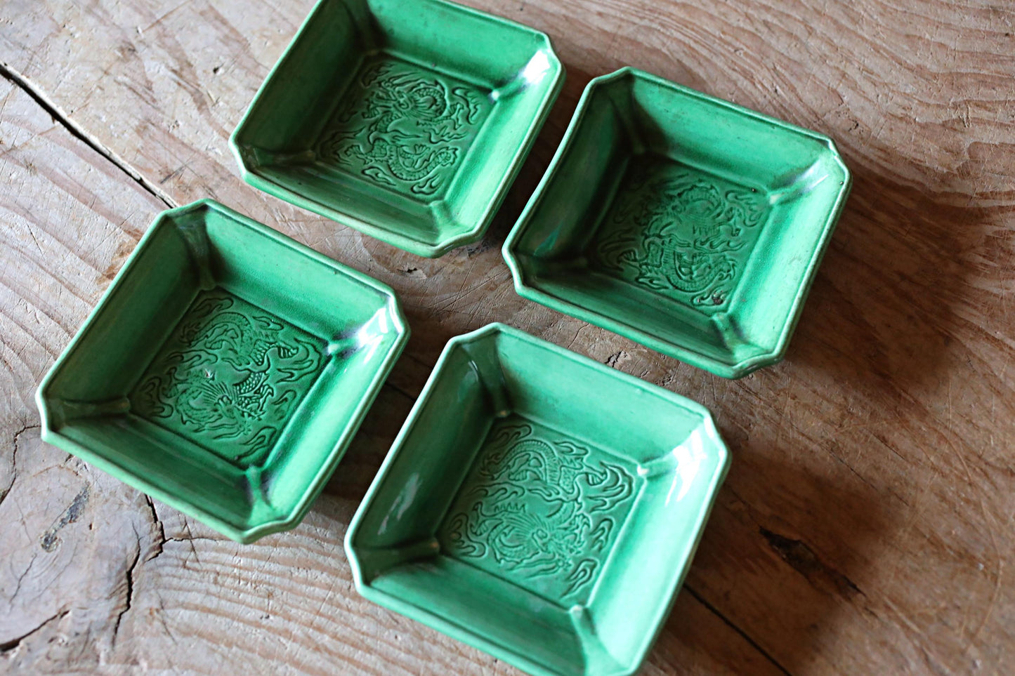 Four square green ceramic dishes with intricate dragon designs are arranged on a wooden surface.