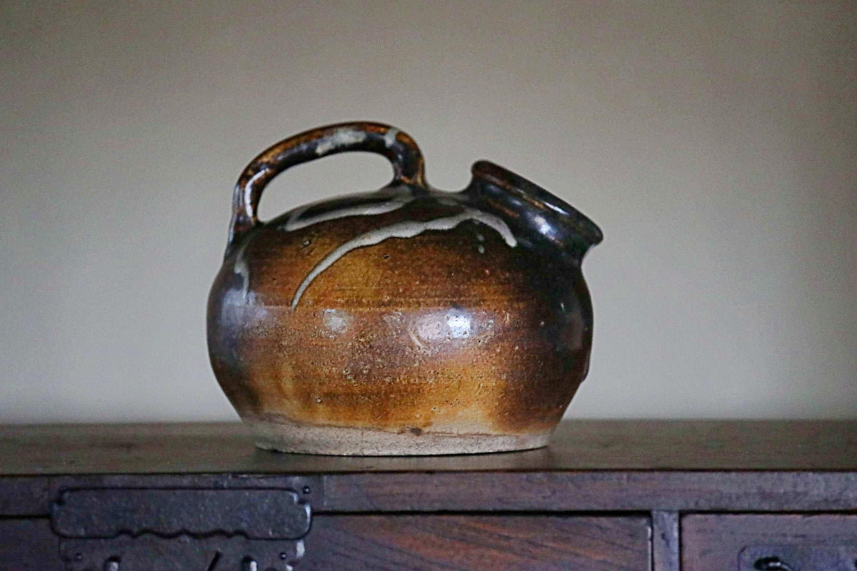 A brown ceramic pitcher with a handle sits on a wooden surface.
