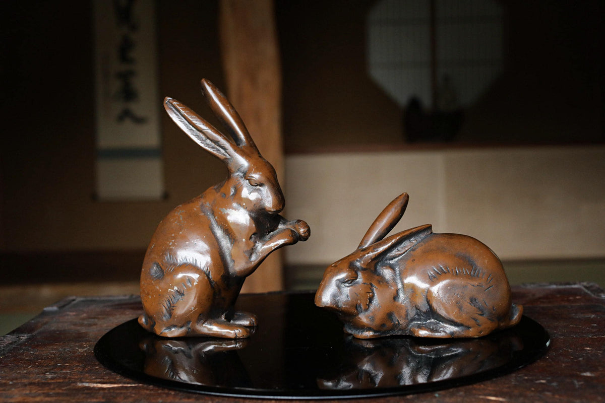 Two bronze rabbit figurines on a black circular tray, one standing and one sitting, in a dimly lit room with a window and a sign with Japanese characters in the background.