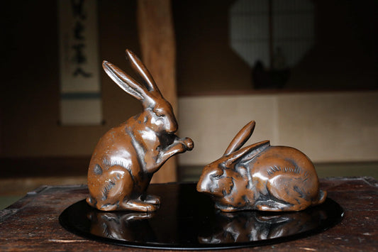 Two bronze rabbit figurines on a black circular tray, one standing and one sitting, in a dimly lit room with a window and a sign with Japanese characters in the background.