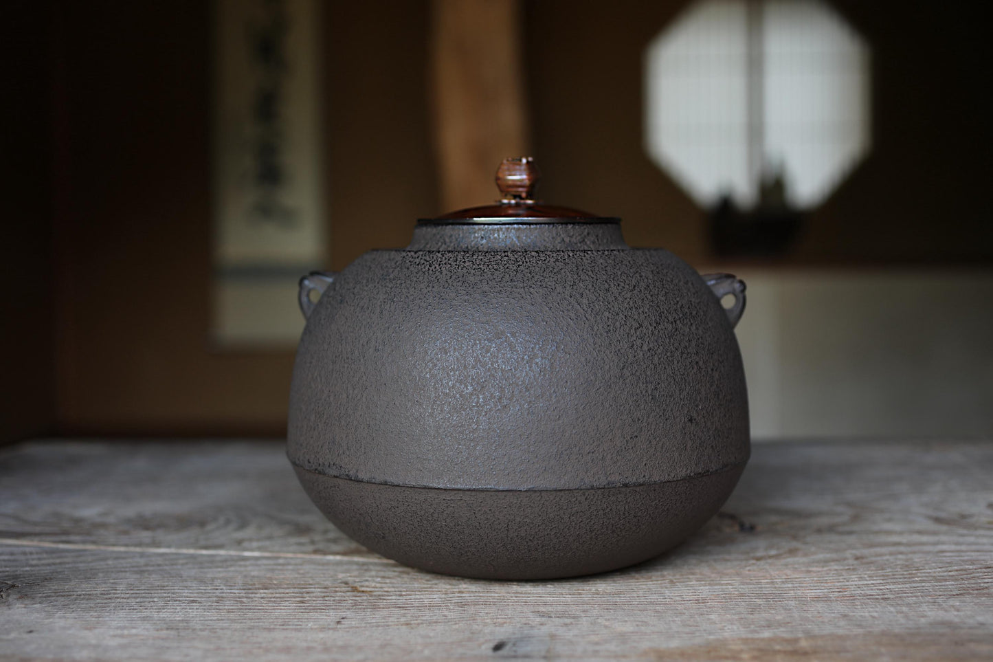 A black ceramic teapot with a wooden lid sits on a wooden surface, with a blurred background suggesting an indoor setting.