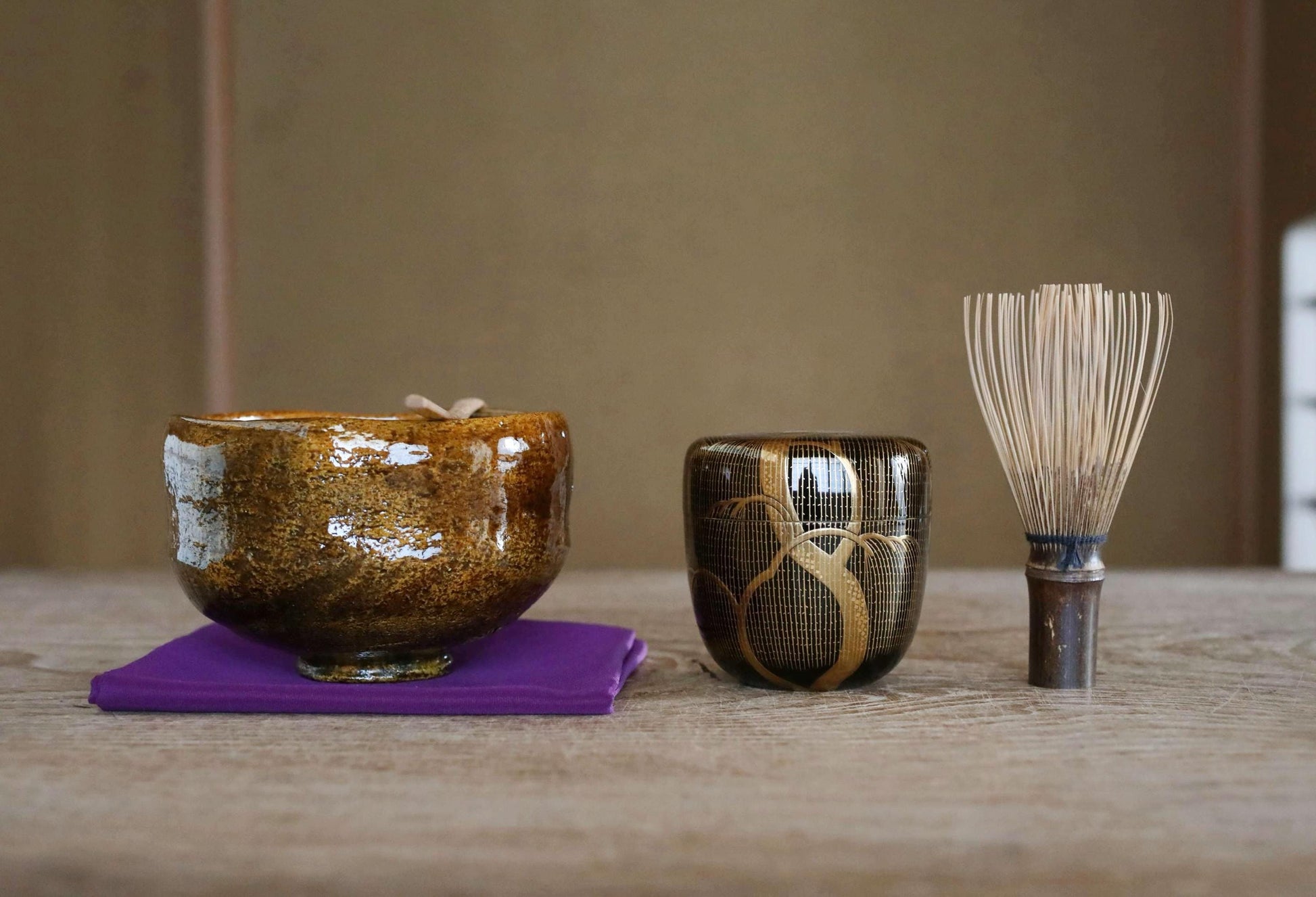 a wooden table with three objects placed on it: a brown ceramic bowl, a black ceramic cup with gold designs, and a wooden whisk.