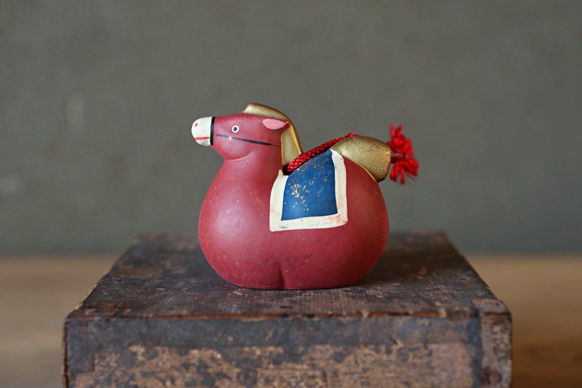 A red toy horse with a blue saddle and red tassel is sitting on a wooden crate against a gray background.