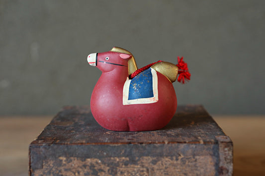 A red toy horse with a blue saddle and red tassel is sitting on a wooden crate against a gray background.