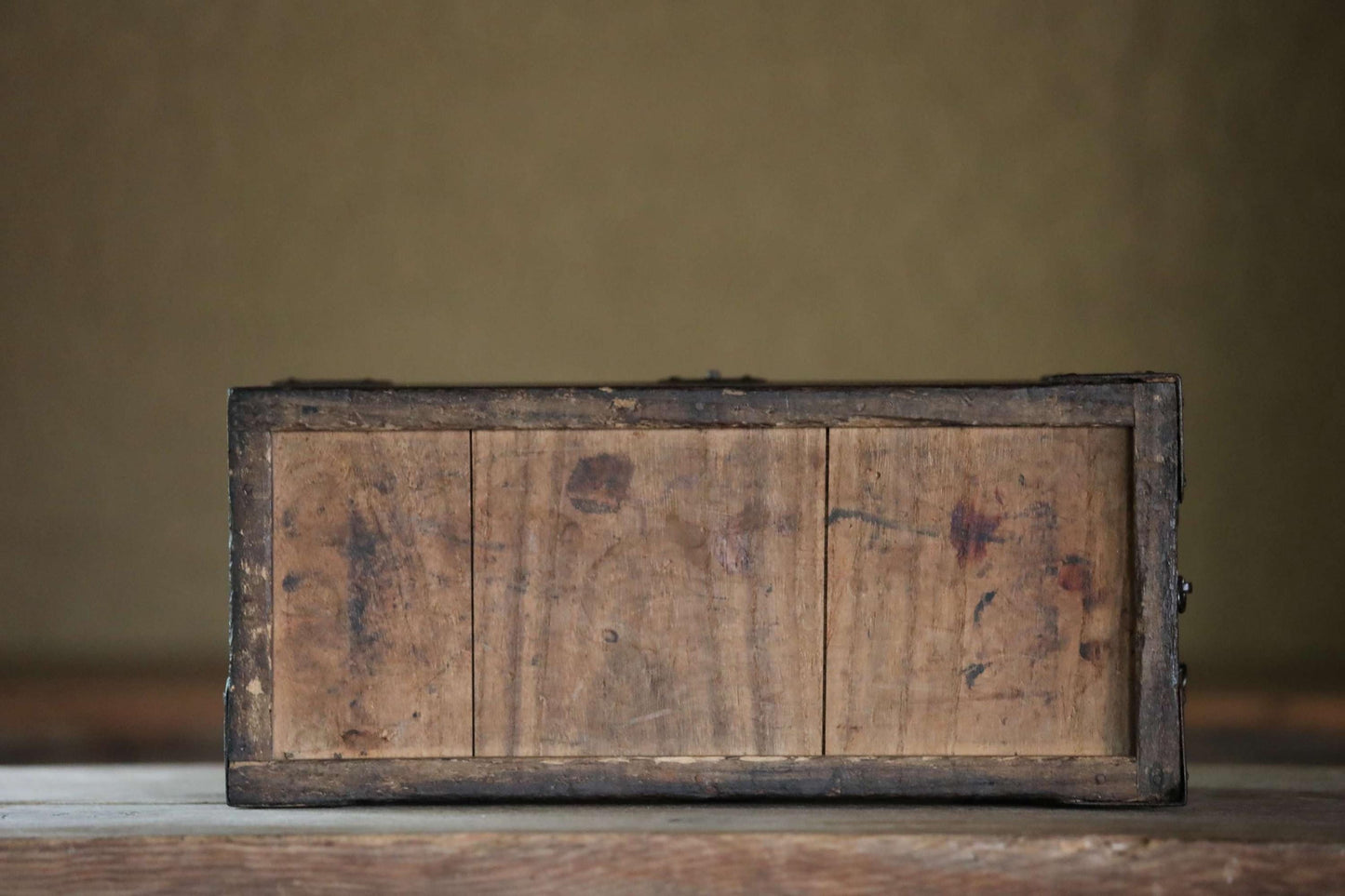 An old, weathered wooden box with a dark finish sits on a wooden surface against a plain background.