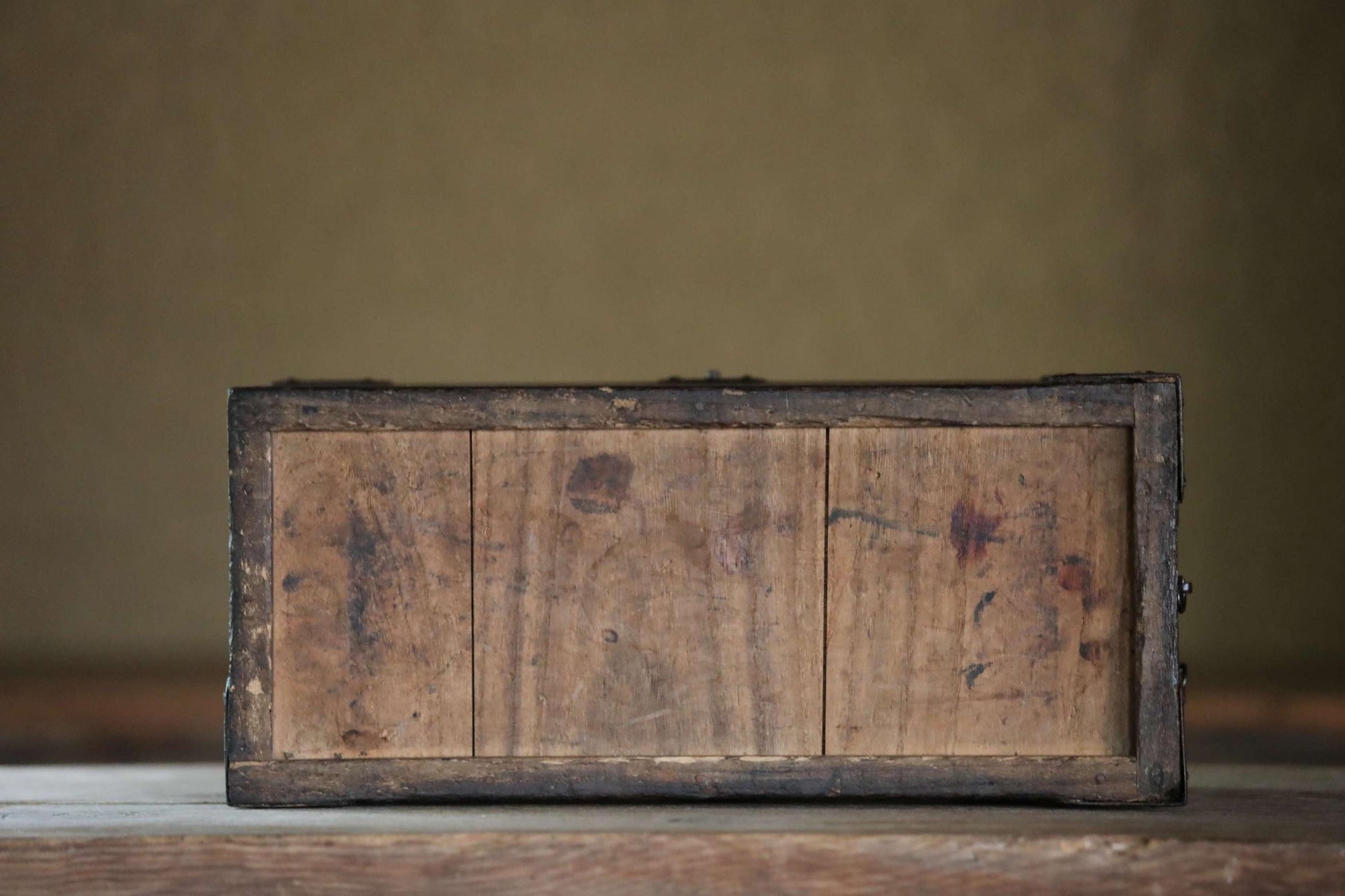 An old, weathered wooden box with a dark finish sits on a wooden surface against a plain background.