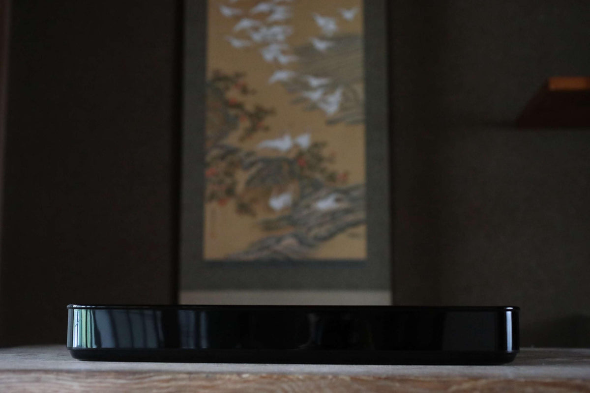 a black rectangular object placed on a wooden surface, with a blurred background that includes a painting or artwork.