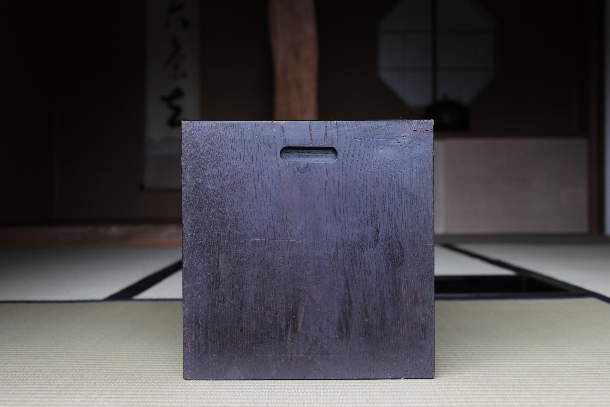A dark wooden box with a hole in the center, placed on a tatami mat in a dimly lit room with a traditional Japanese-style wall in the background.