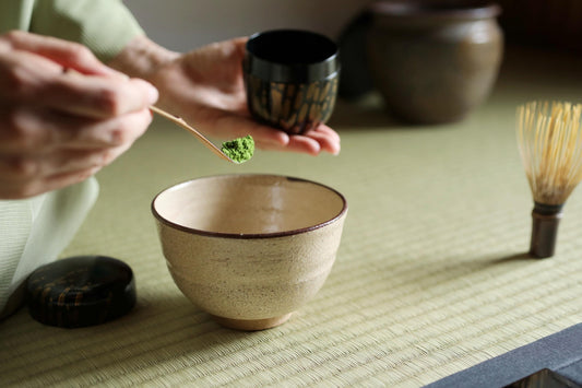A person is preparing a cup of matcha tea, using a whisk to mix the matcha powder into a small bowl.