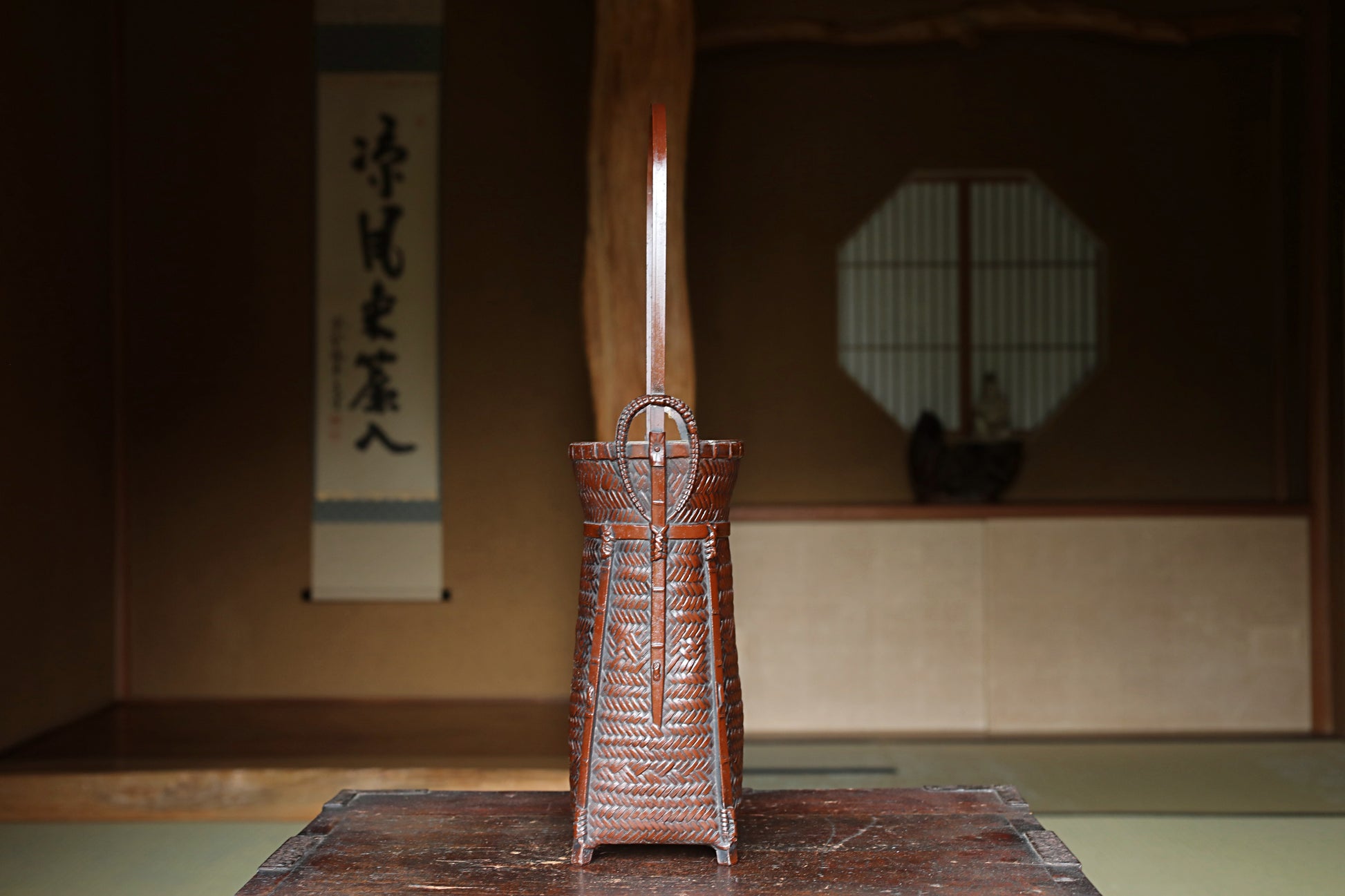 A wooden basket with a curved handle, placed on a wooden surface, with a traditional Japanese-style room in the background.