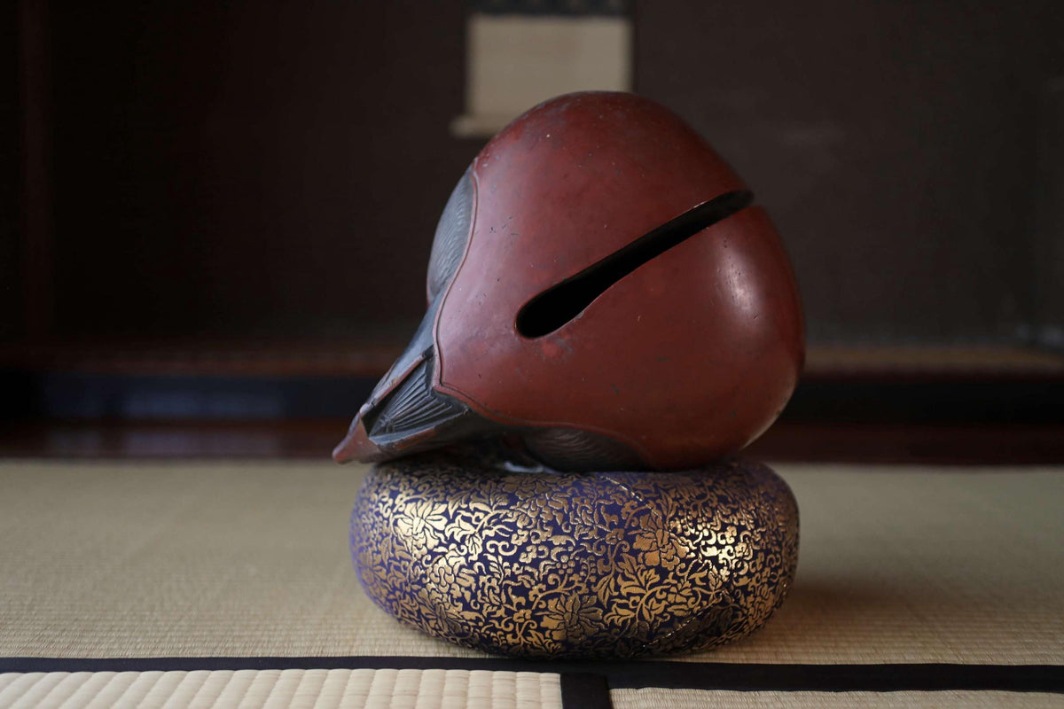 A red and black object, possibly a sculpture or vase, is placed on a blue and gold decorative item, which appears to be a small ceramic bowl or plate, on a woven mat or rug.