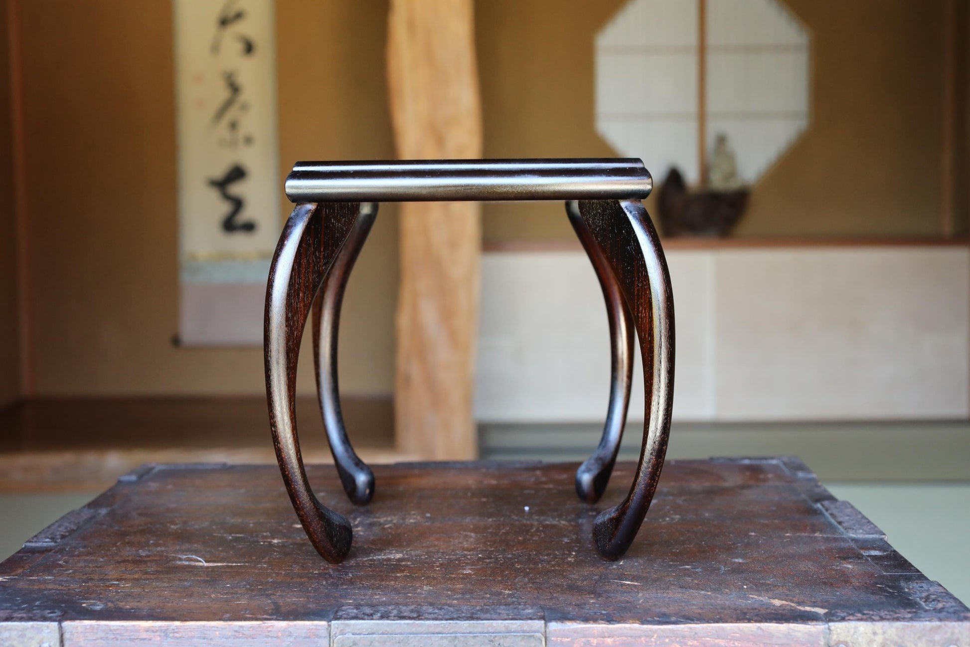 A wooden table with a curved top and legs, placed in an interior setting with a traditional Japanese-style wall in the background.