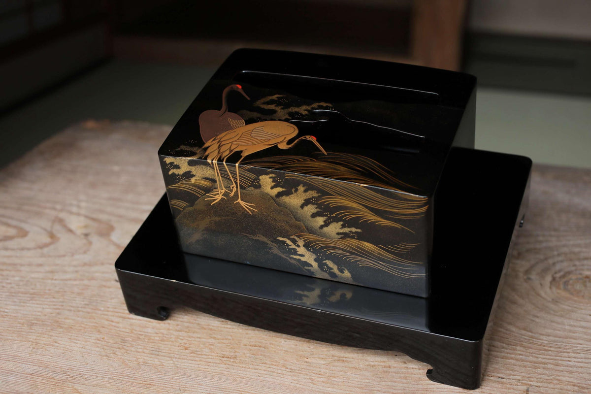 A black box with a gold design depicting two cranes on a wooden surface.
