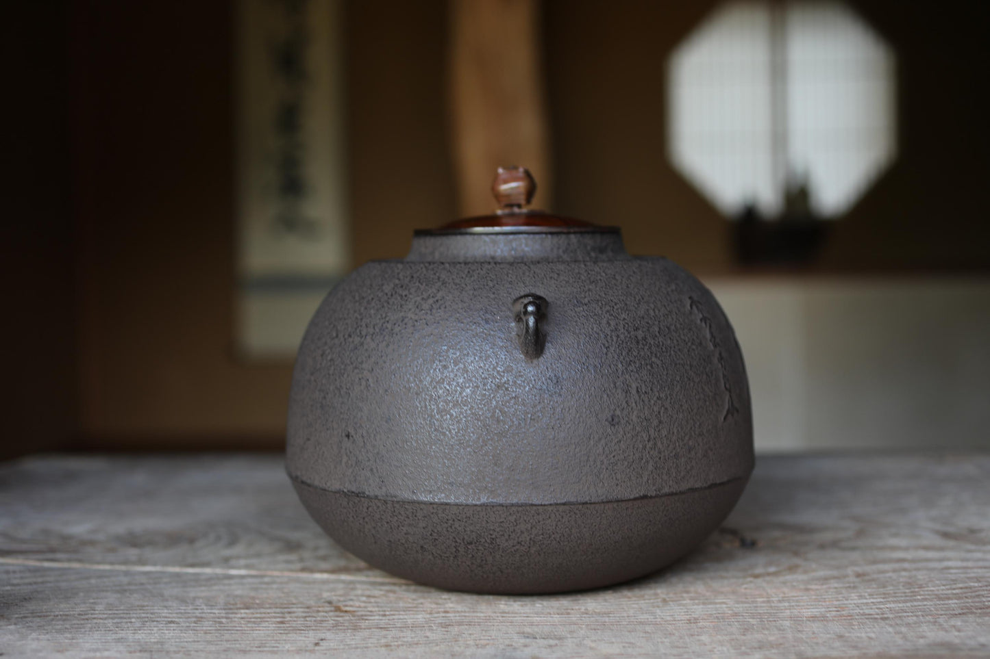 A black, round tea kettle with a brown lid sits on a wooden surface, with a blurred background that includes a window and a wall with Japanese characters.