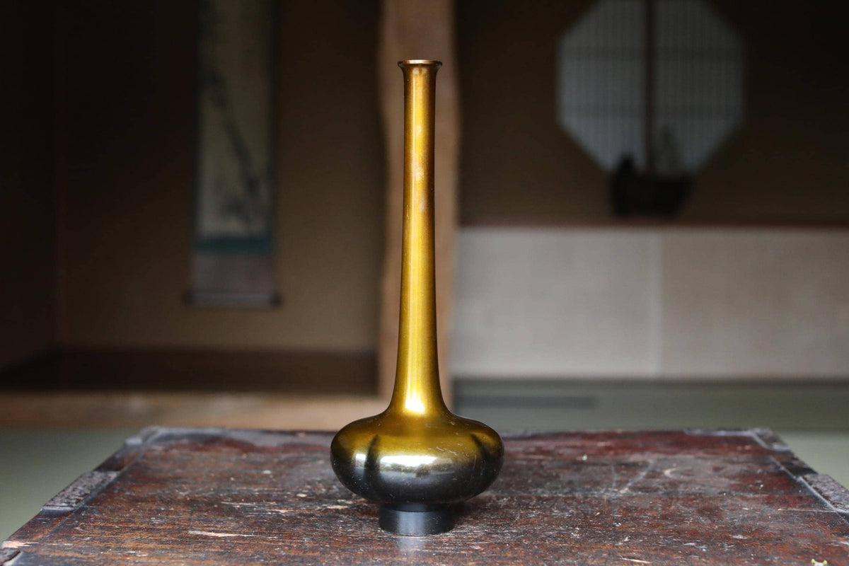 A golden vase with a long, slender shape sits on a wooden surface, with a blurred background that includes a window and a wall.