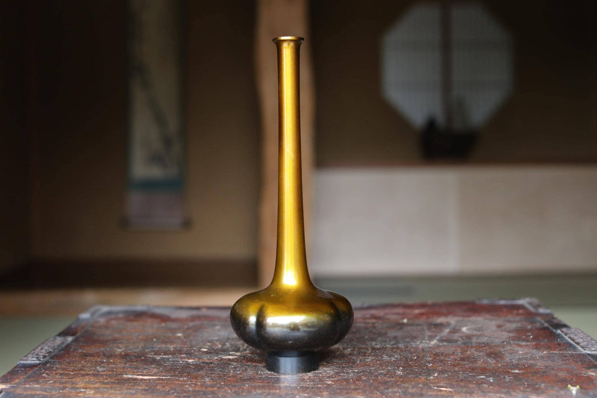 A golden vase with a long, slender shape sits on a wooden surface, with a blurred background suggesting an indoor setting.