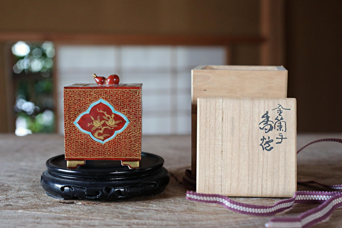 a wooden box with a red and gold design, placed on a black stand, and a wooden box with Japanese characters on it, both sitting on a wooden surface.