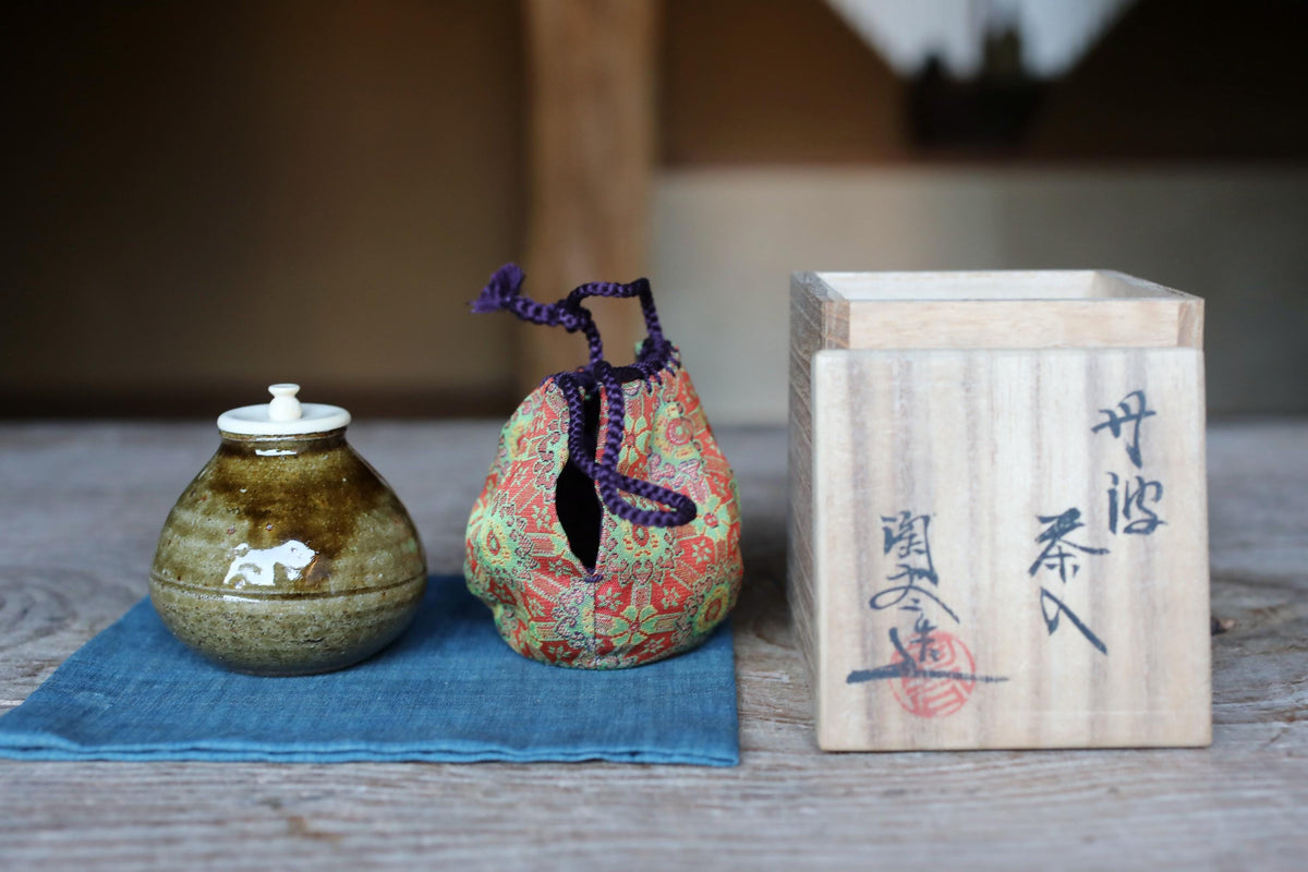 a ceramic jar with a white lid and a colorful cloth bag with a purple handle, both placed on a blue cloth on a wooden surface. There is also a wooden box with Japanese characters on it.
